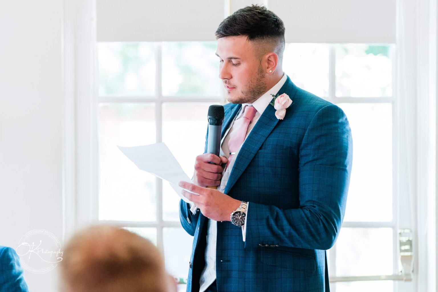Warwick House Wedding Photography A man in a blue suit, pink tie, and boutonniere is giving a speech while holding a microphone and a piece of paper during a wedding at Warwick House.