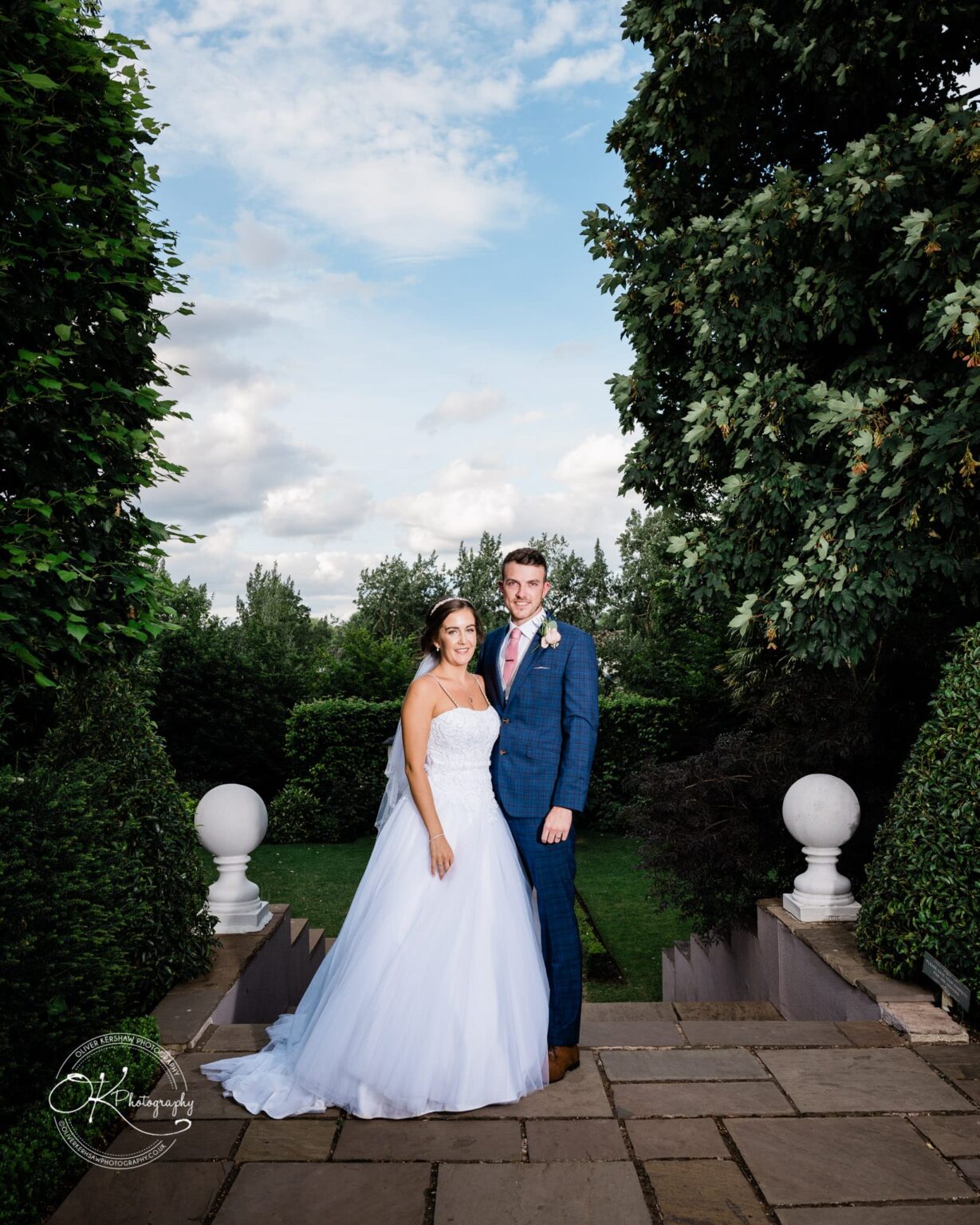 Warwick House Wedding Photography A bride and groom stand together in formal wedding attire, posing outdoors surrounded by greenery at an elegant venue.