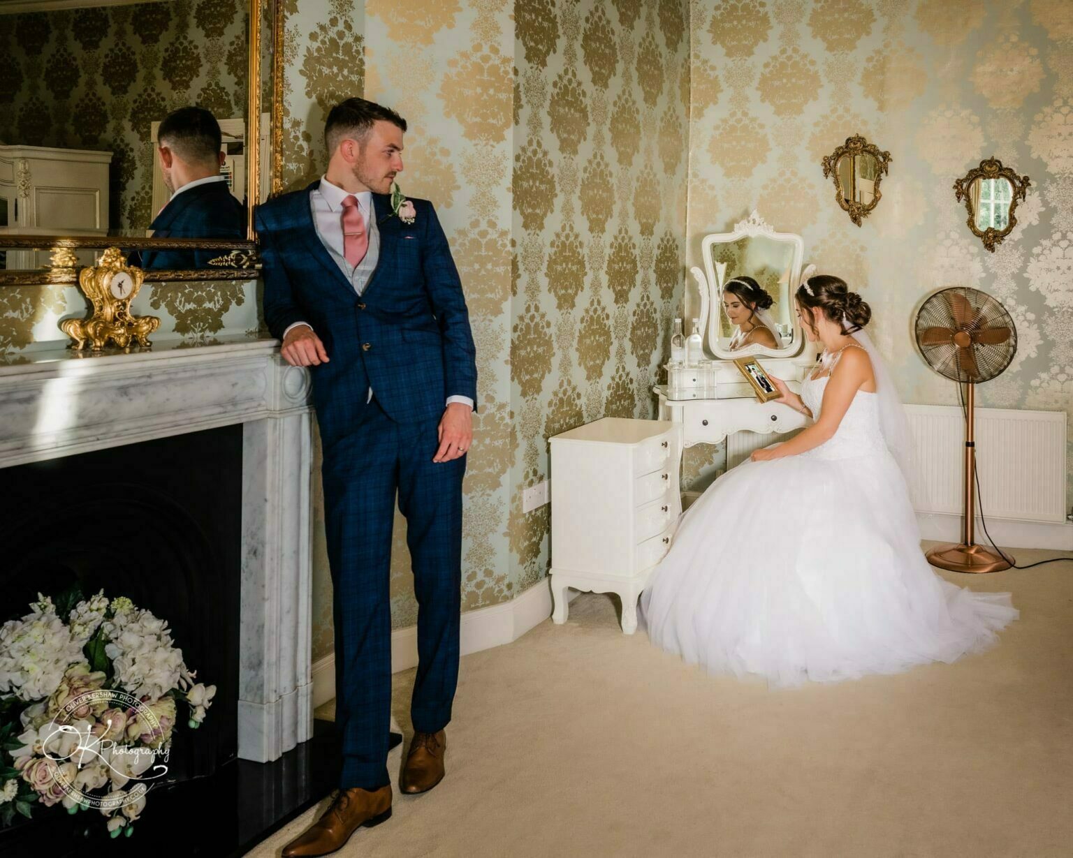 Warwick House Wedding Photography A bride in a white gown sits at a vintage dressing table, while the groom in a blue suit leans against a marble fireplace in an elegant, ornate room.