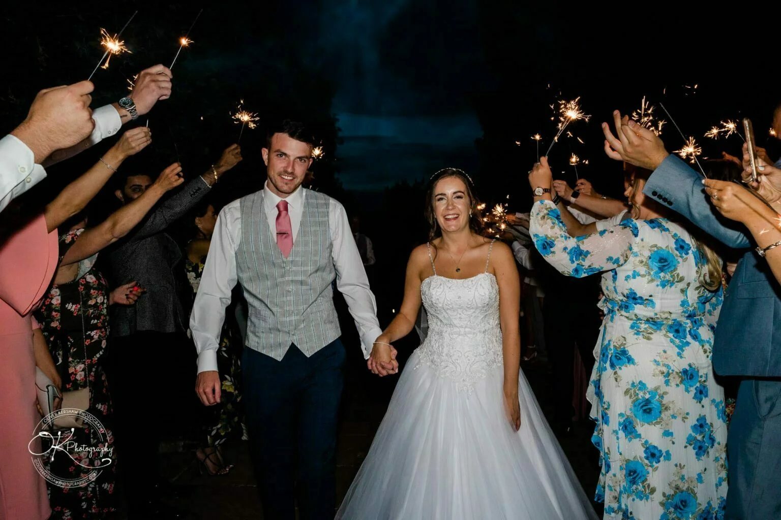 Warwick House Wedding Photography A bride and groom holding hands and smiling while walking through a group of guests holding sparklers at night.