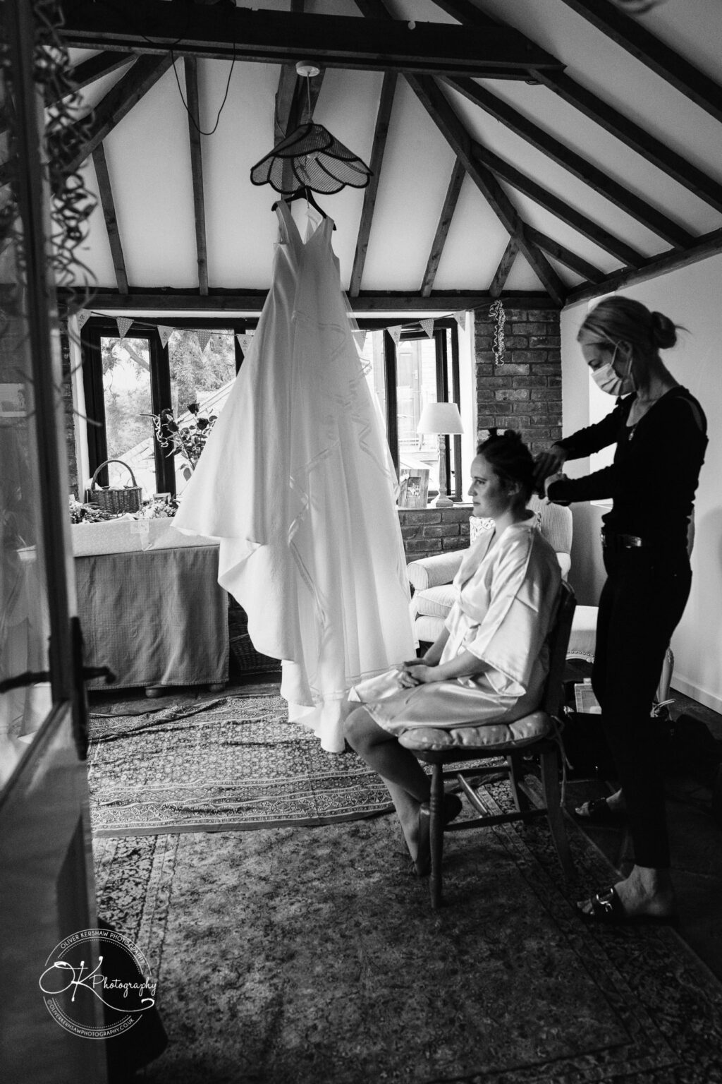 Wedding photography St Michael & All Angels Church A woman having her hair styled while sitting in a chair in a rustic room with exposed beams and a wedding dress hanging from the ceiling.