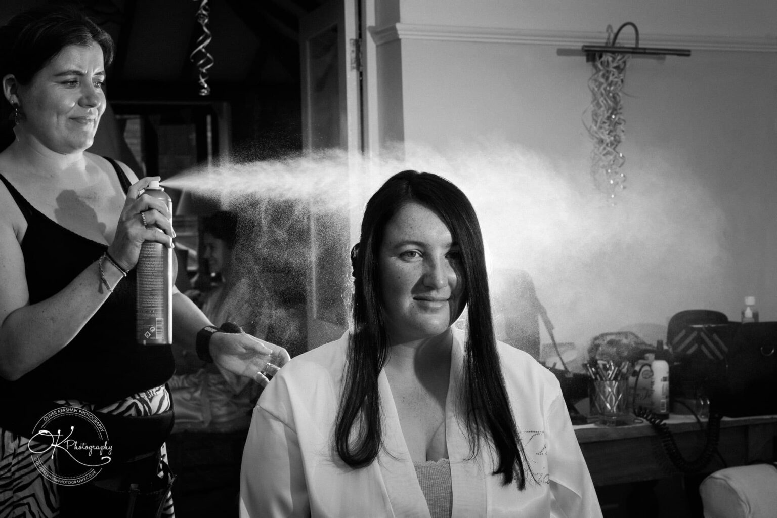 Wedding photography St Michael & All Angels Church A woman getting her hair styled with hairspray by another woman, in a black and white photograph.