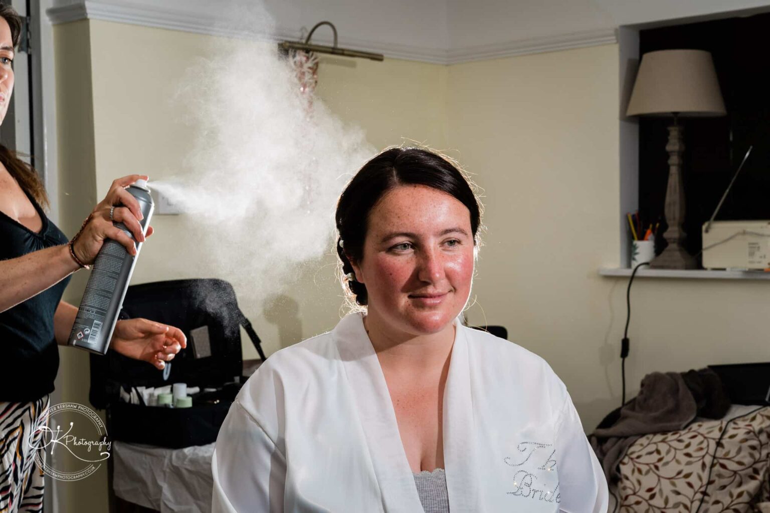 Wedding photography St Michael & All Angels Church A woman in a white robe having her hair sprayed with hairspray in a well-lit room.