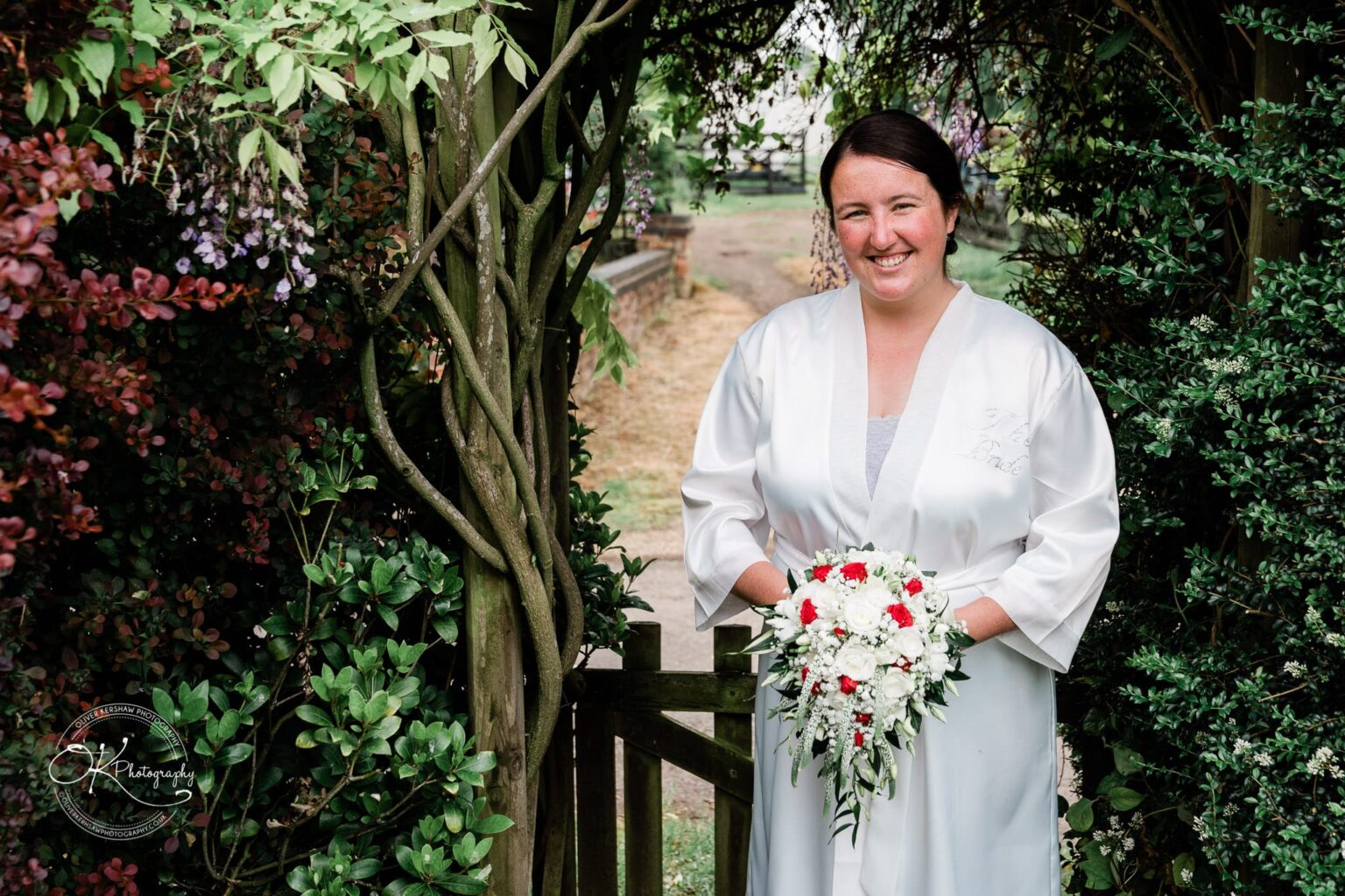 Wedding photography St Michael & All Angels Church A woman in a white robe holding a bouquet of red and white flowers, standing in a garden archway.