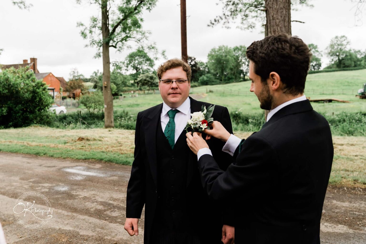 Wedding photography St Michael & All Angels Church Two men in formal wear, one adjusting a boutonnière on the other's suit, in an outdoor setting near a church with greenery and trees in the background.