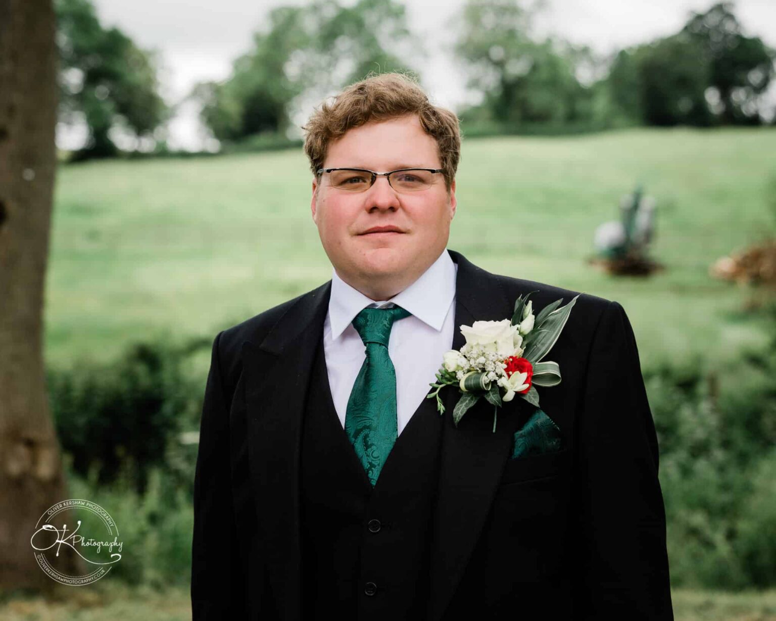 Wedding photography St Michael & All Angels Church A man wearing a black suit, white shirt, and green tie stands outdoors with a boutonnière pinned to his lapel. The background features a green, grassy area with trees.