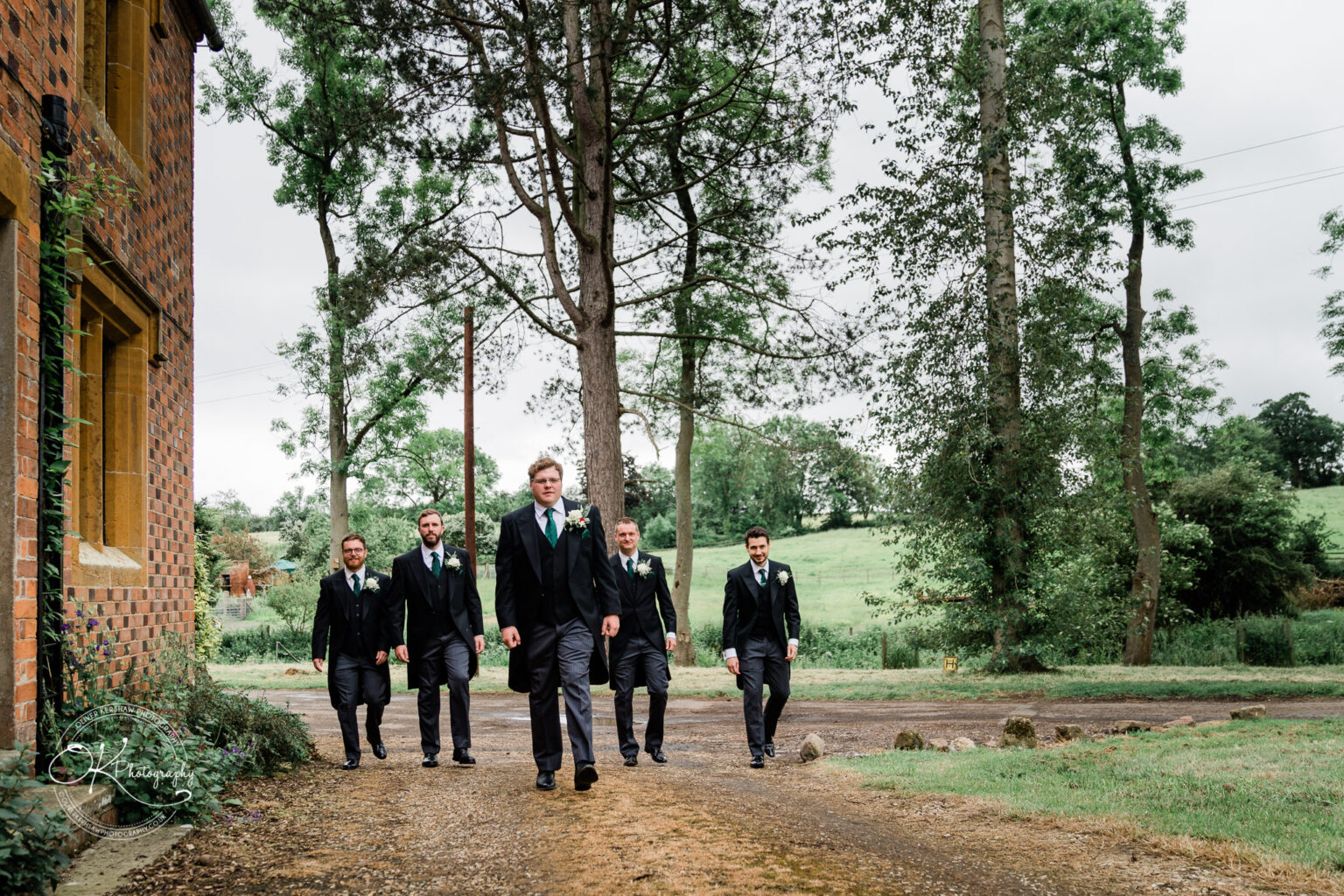 Wedding photography St Michael & All Angels Church Five men in suits walking outside near a brick building and trees.