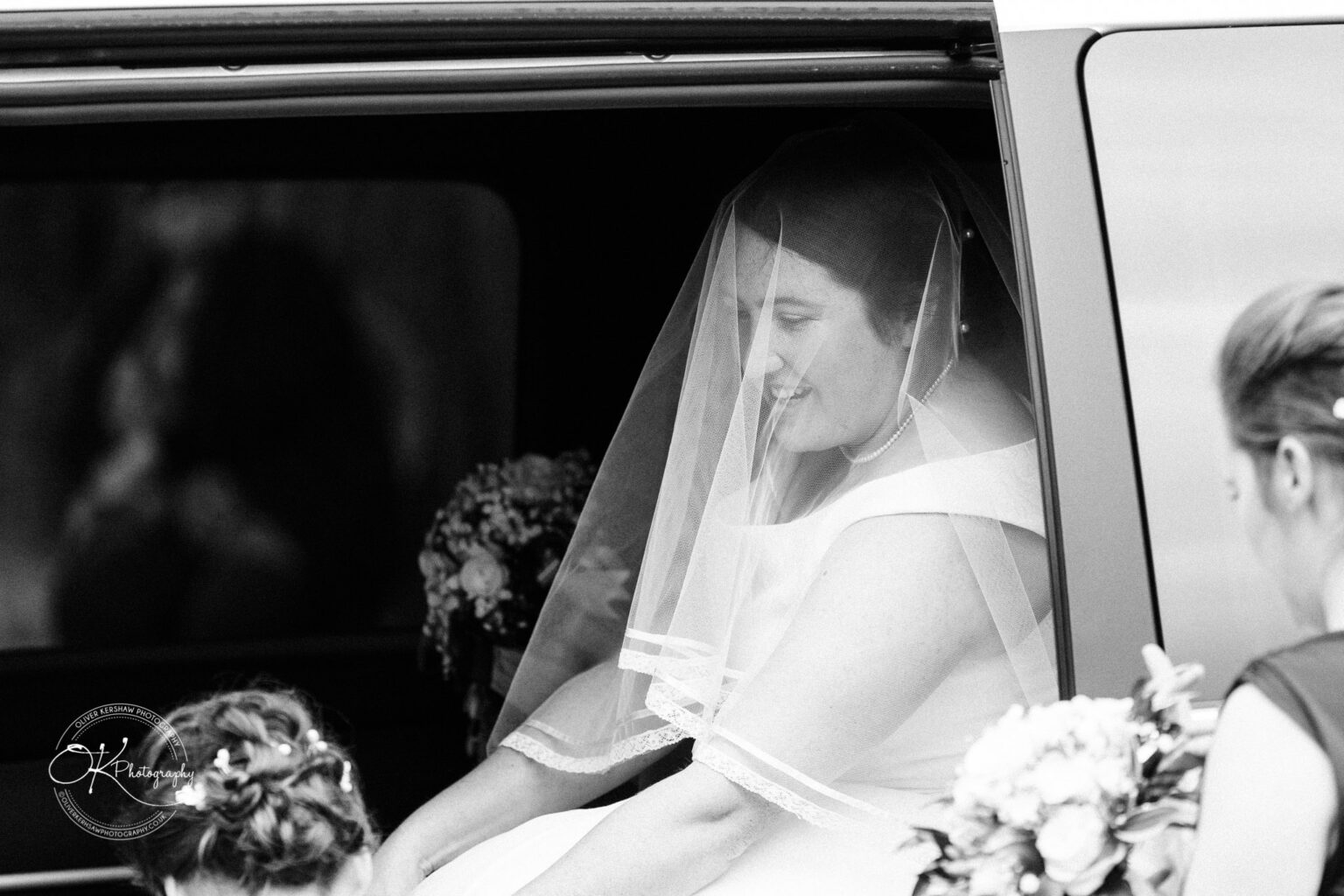 Wedding photography St Michael & All Angels Church Black and white photo of a smiling bride wearing a veil, in the doorway of a vehicle, with a floral bouquet and an attendant visible.