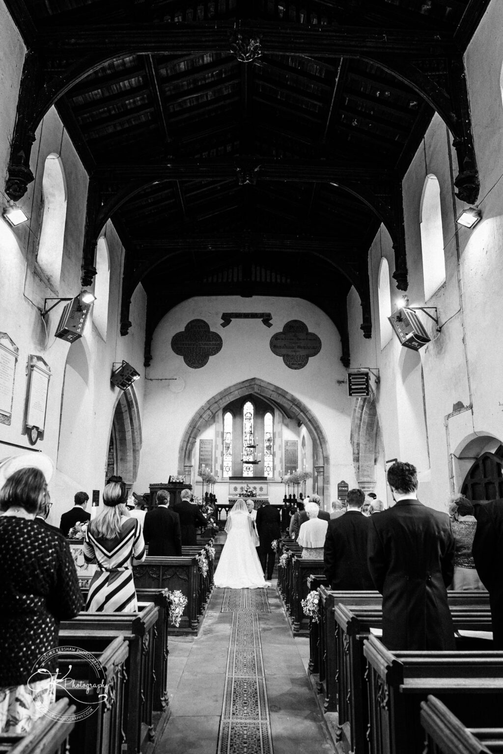 Wedding photography St Michael & All Angels Church A wedding ceremony taking place inside a traditional church with a high wooden ceiling, arched windows, and pews filled with guests.