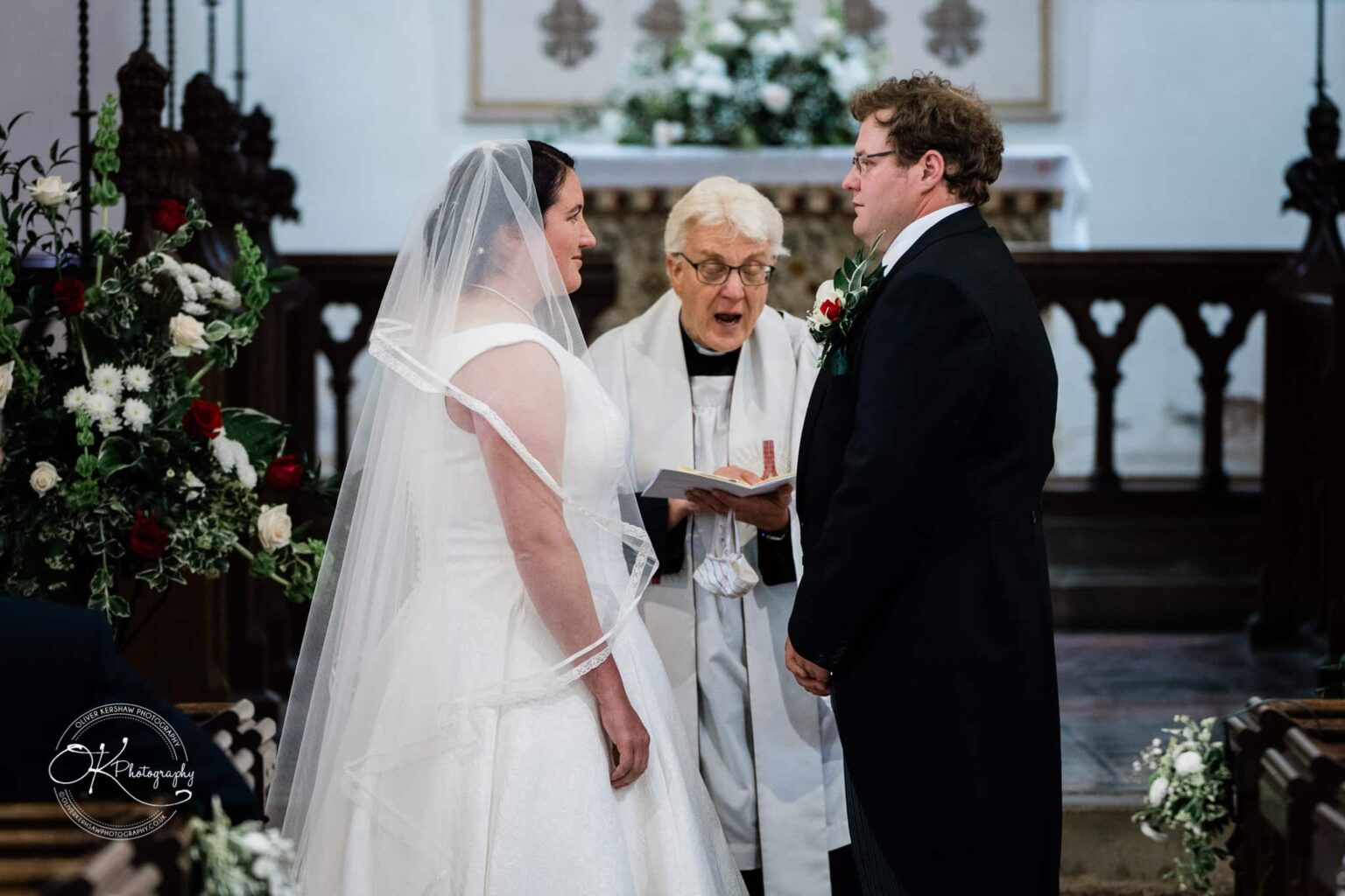Wedding photography St Michael & All Angels Church A wedding ceremony in progress at St Michael's Church, with a bride in a white dress and veil and a groom in a suit, standing before a priest.