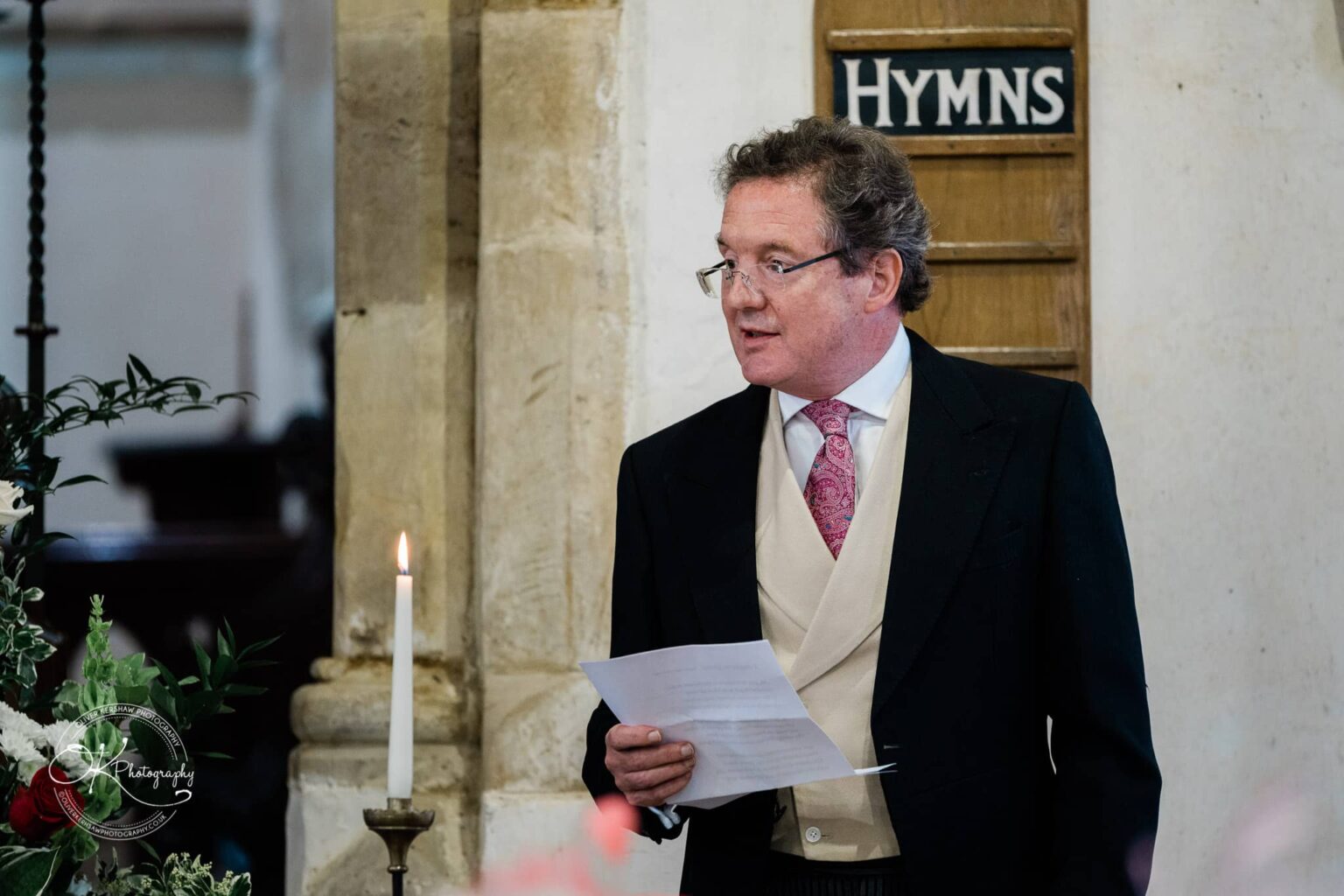 Wedding photography St Michael & All Angels Church A man in formal attire speaks inside a church, standing near a lit candle and floral arrangement, with a hymns board in the background.