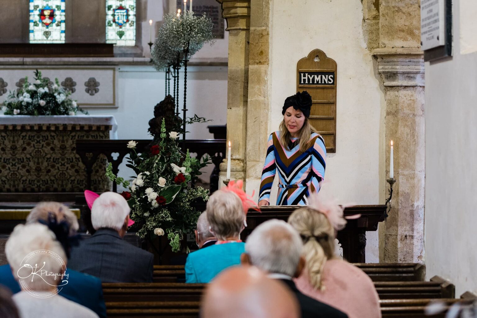 Wedding photography St Michael & All Angels Church A woman in colourful attire reads at a lectern inside a church, with floral arrangements and stained glass windows in the background. Congregants are seated in pews, facing forward.
