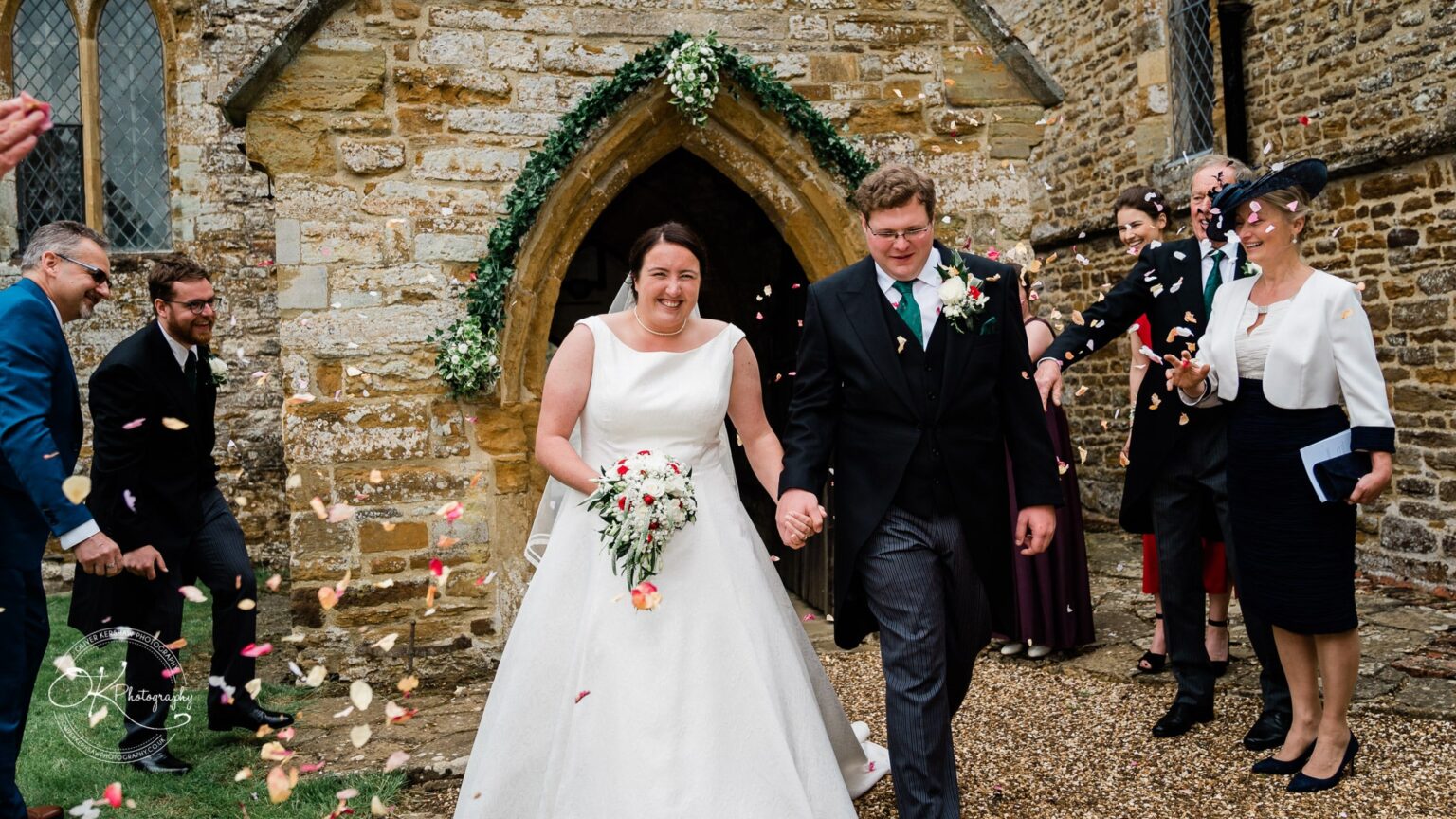 Wedding photography St Michael & All Angels Church Bride and groom smiling and holding hands while exiting St Michael's Church, surrounded by guests throwing flower petals.