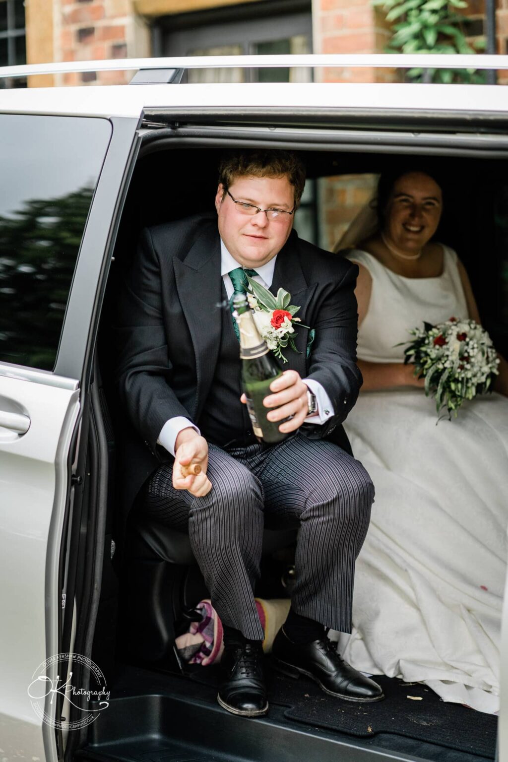Wedding photography St Michael & All Angels Church A man dressed in formal wedding attire holding a bottle of champagne, sitting at the edge of a vehicle door, with a woman in a wedding dress holding a bouquet in the background.