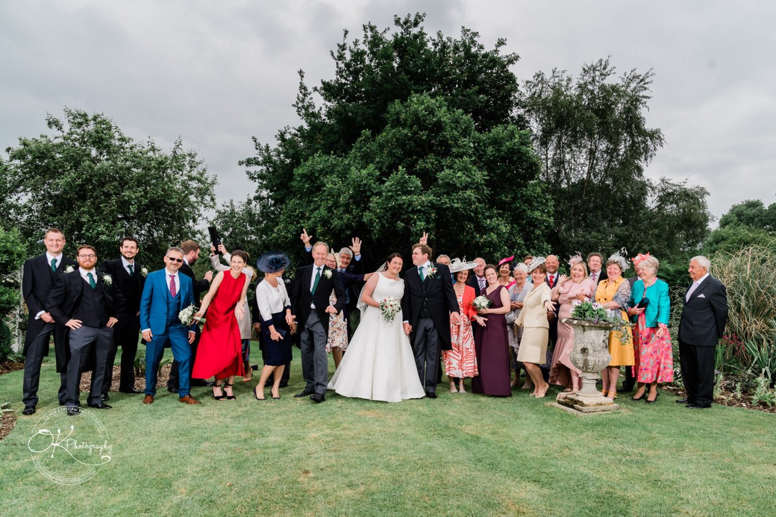 Wedding photography St Michael & All Angels Church A wedding party group photo taken outdoors in front of a large tree, with all participants dressed in formal attire and smiling at the camera.