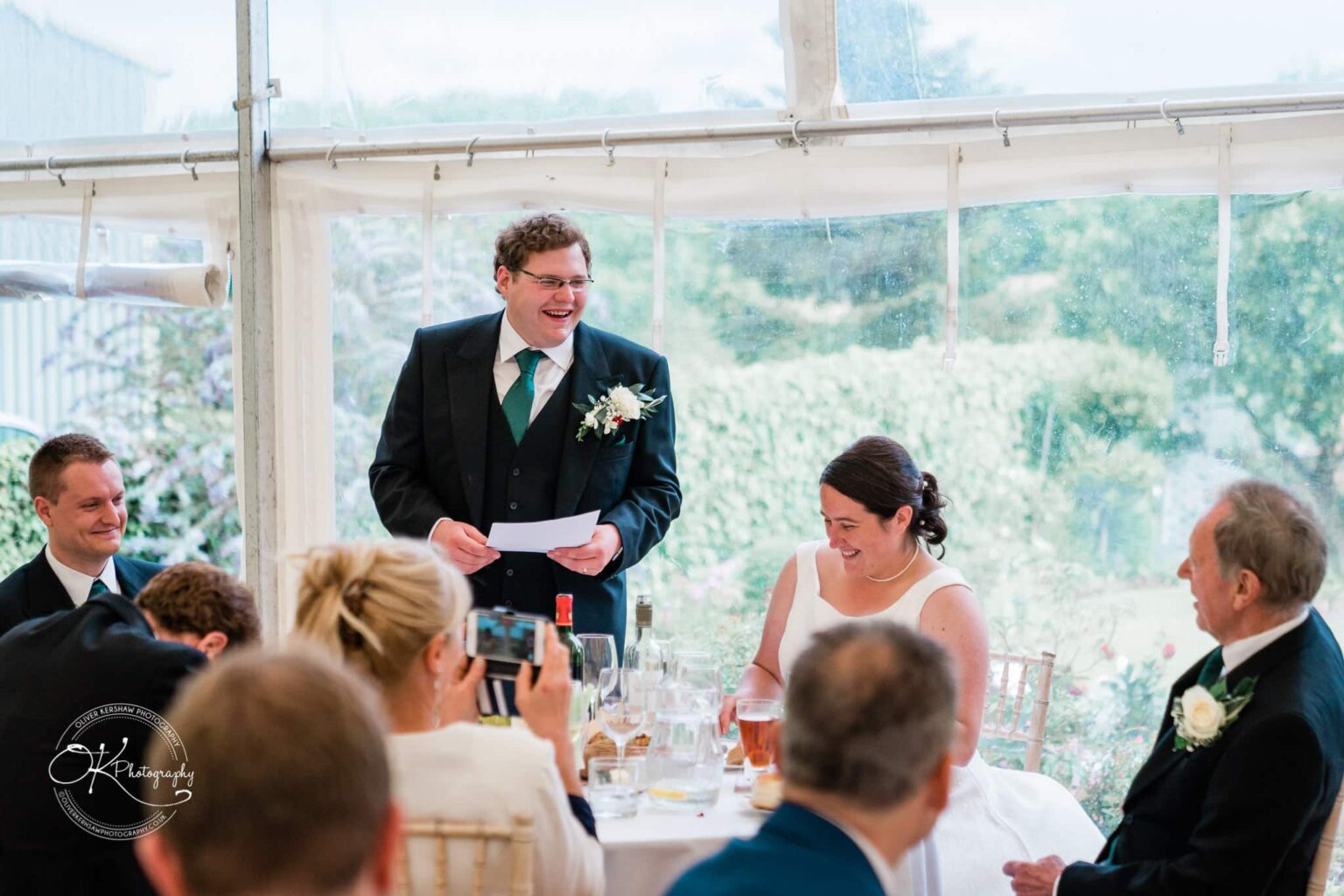 Wedding photography St Michael & All Angels Church A groom giving a speech while standing beside his bride at a wedding reception, with guests seated around them in a marquee.