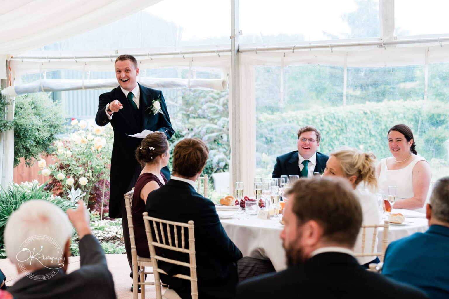 Wedding photography St Michael & All Angels Church A man in a suit giving a speech at a wedding reception while guests seated around a table listen and smile inside a decorated marquee.
