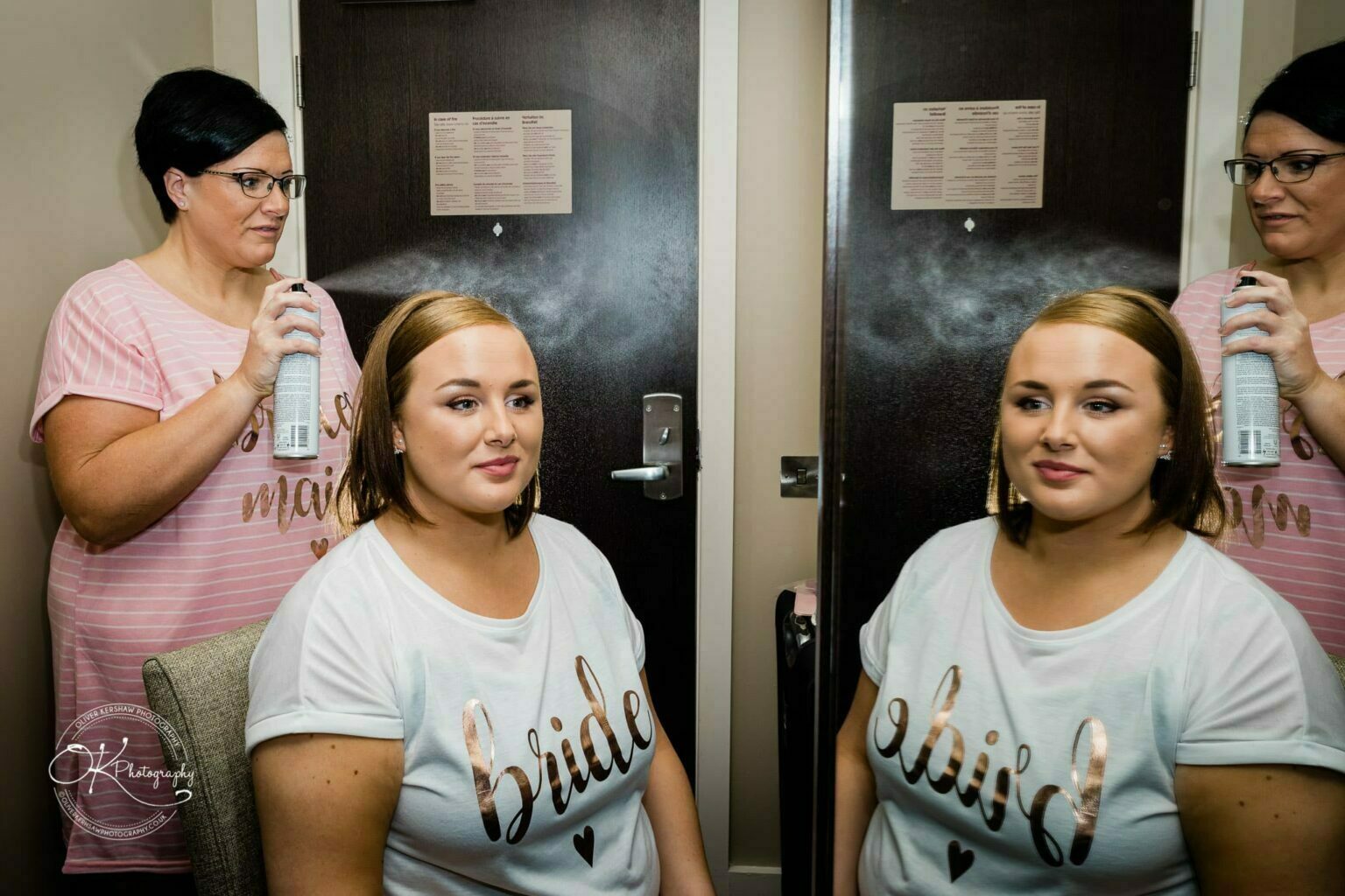 Wedding photography The Embankment A woman wearing a "bride" T-shirt is seated while another woman, wearing a "bridesmaid" T-shirt, sprays her hair with hairspray in front of a mirror.
