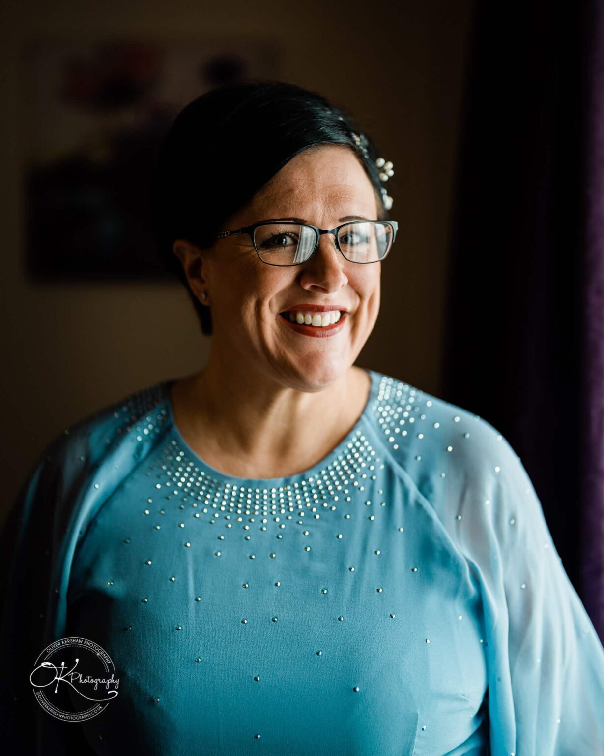 Wedding photography The Embankment A woman wearing glasses and a light blue, beaded top is smiling against a dark background.