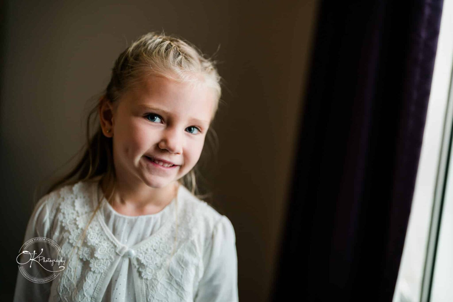 Wedding photography The Embankment A young girl with braided blonde hair and dimples, wearing a white lace top, standing and smiling near a window.