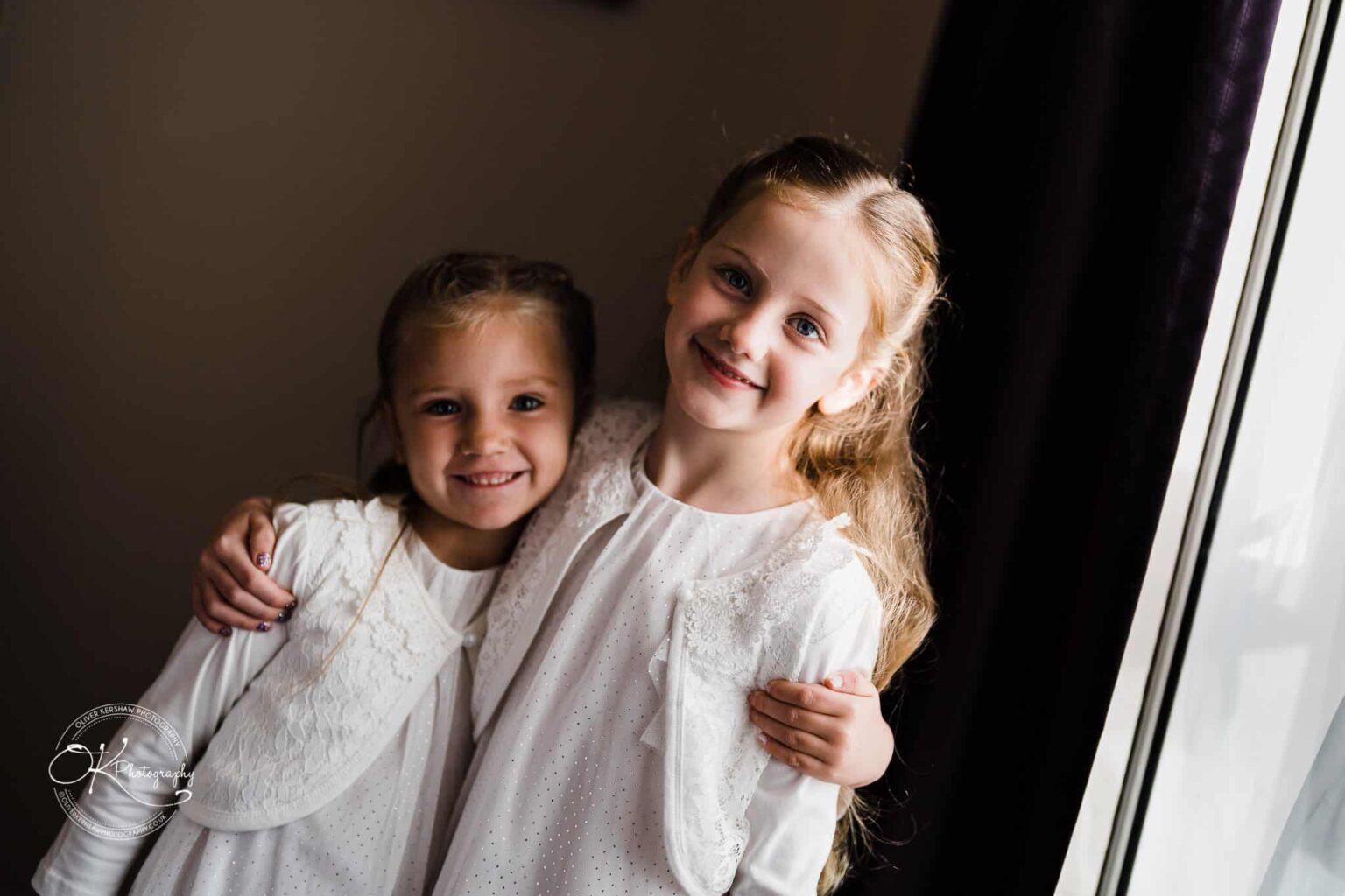 Wedding photography The Embankment Two young girls smiling and embracing, standing near a window with natural light illuminating them.