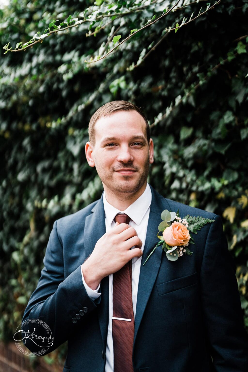 Wedding photography The Embankment Man in a dark suit and red tie, with an orange boutonnière, standing in front of ivy-covered wall.