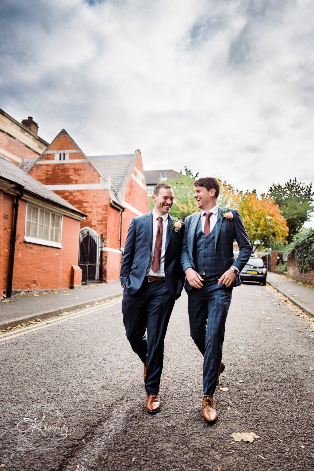 Wedding photography The Embankment Two men in suits walking down a street, smiling at each other, with a red brick building and cloudy sky in the background.