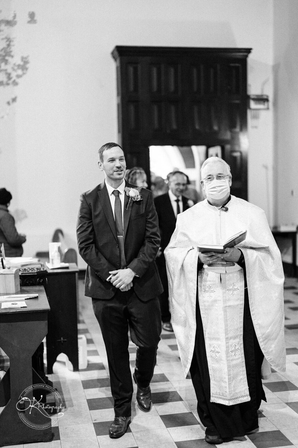 Wedding photography The Embankment A groom in a suit and tie stands next to a priest holding a book, with people in the background at a church ceremony. The priest is wearing a mask.