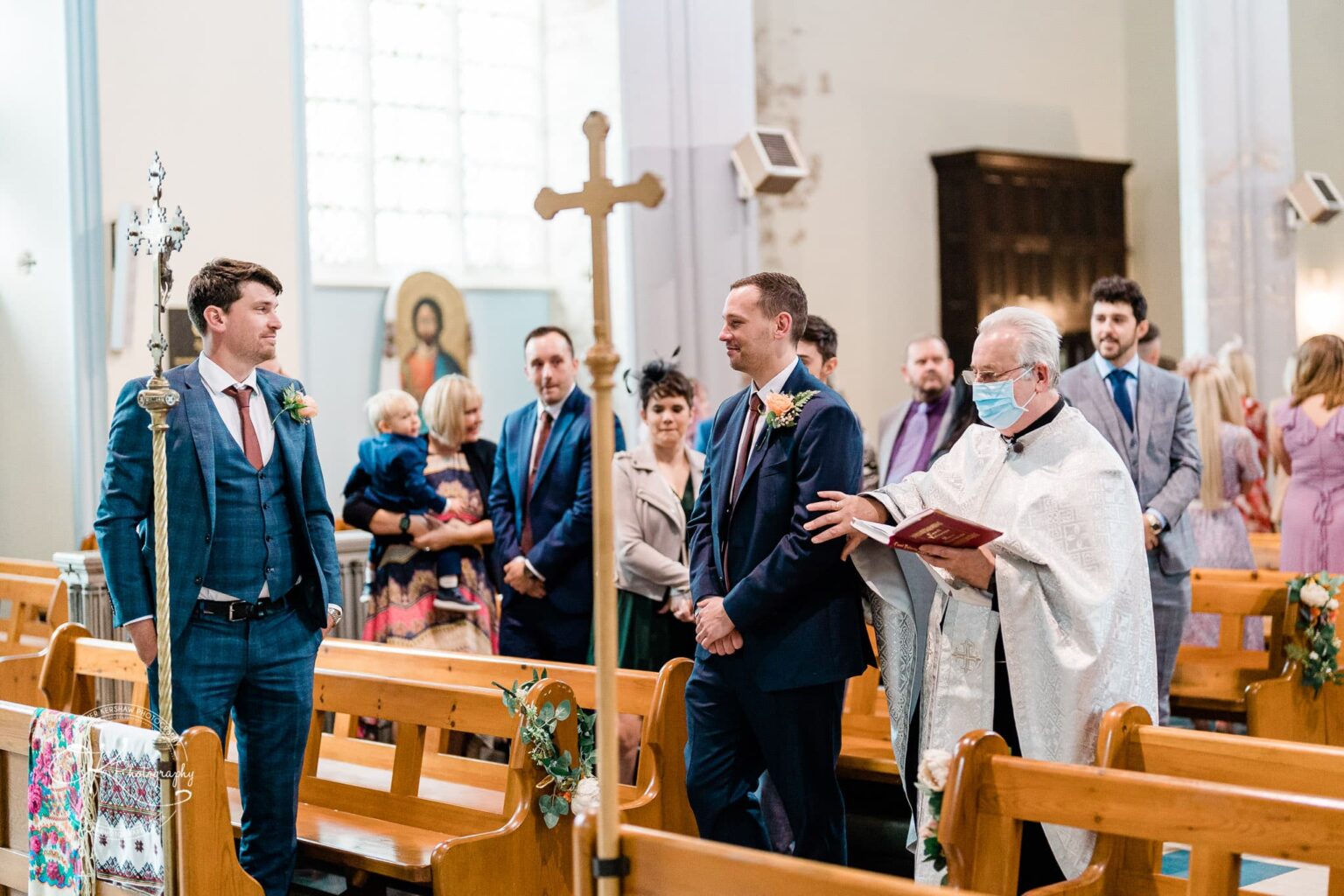 Wedding photography The Embankment A priest wearing a mask and holding a book leads a wedding ceremony in a church, with the groom and attendees standing in the aisle.