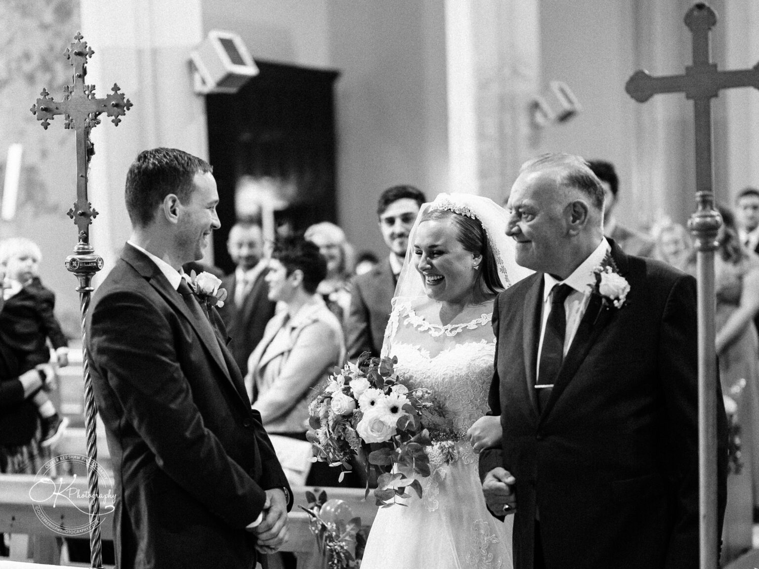 Wedding photography The Embankment Black and white photo of a bride and groom, with the bride accompanied by an older man, smiling at each other during a wedding ceremony in a church.