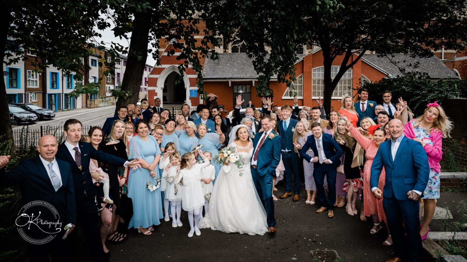 Wedding photography The Embankment A large group wedding photo with the bride and groom surrounded by guests outside a red brick building and trees in the background.