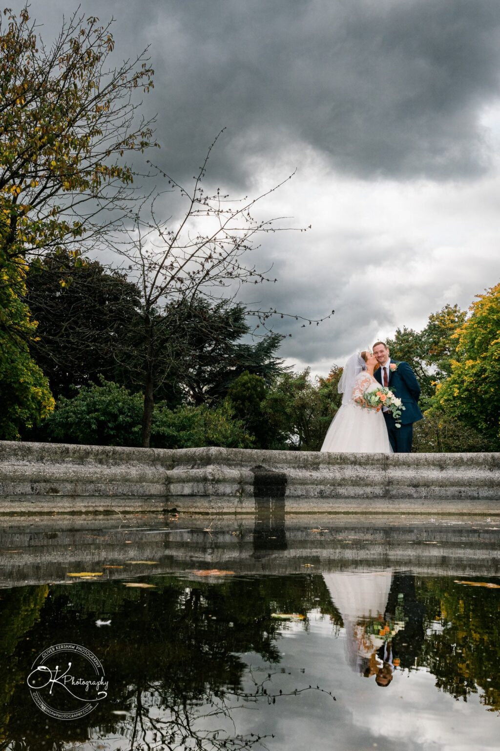 Wedding photography The Embankment A bride and groom in wedding attire standing beside a reflective water surface, with trees and a cloudy sky in the background.