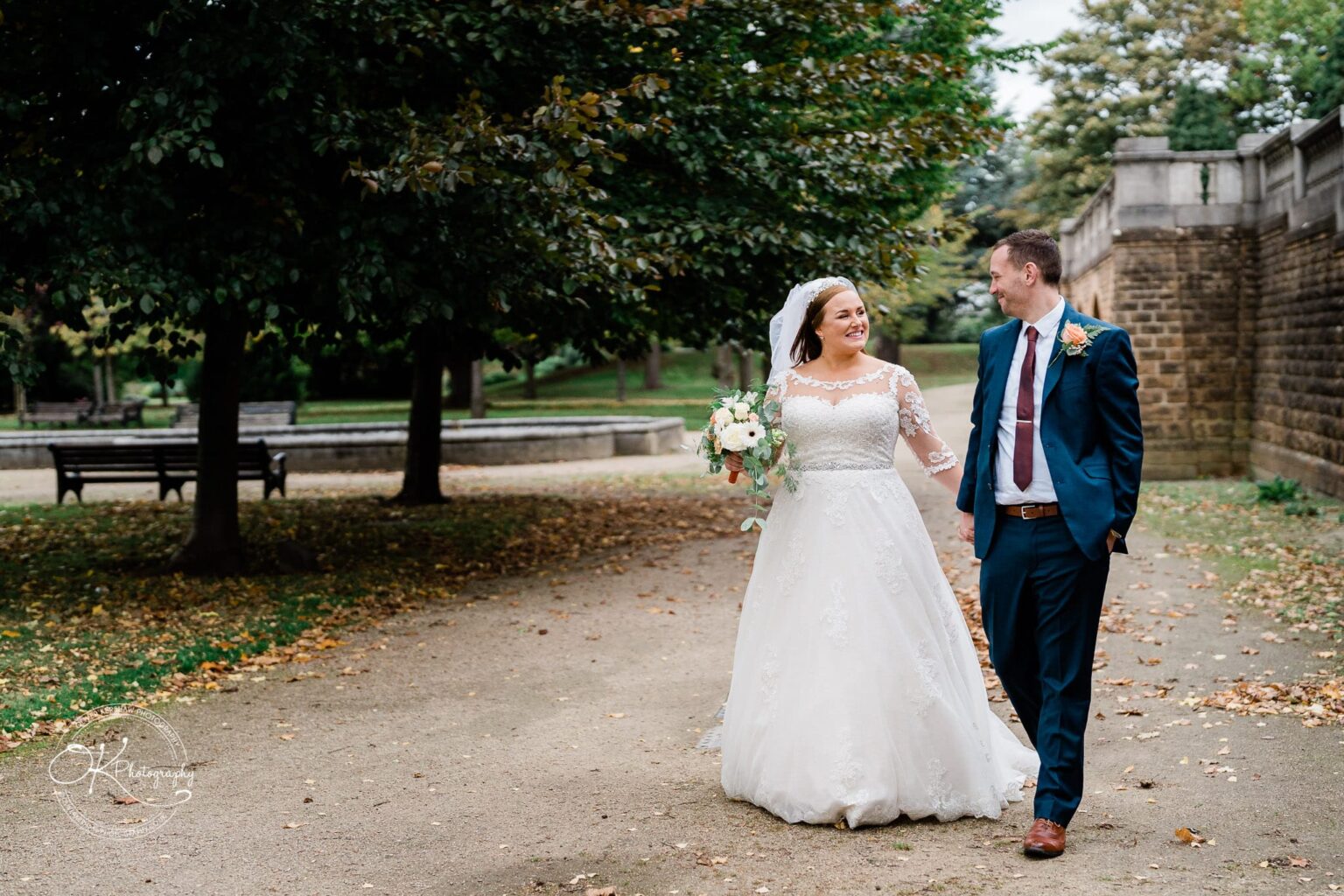 Wedding photography The Embankment Bride and groom walking through a park, with the bride wearing a white lace dress and holding a bouquet.