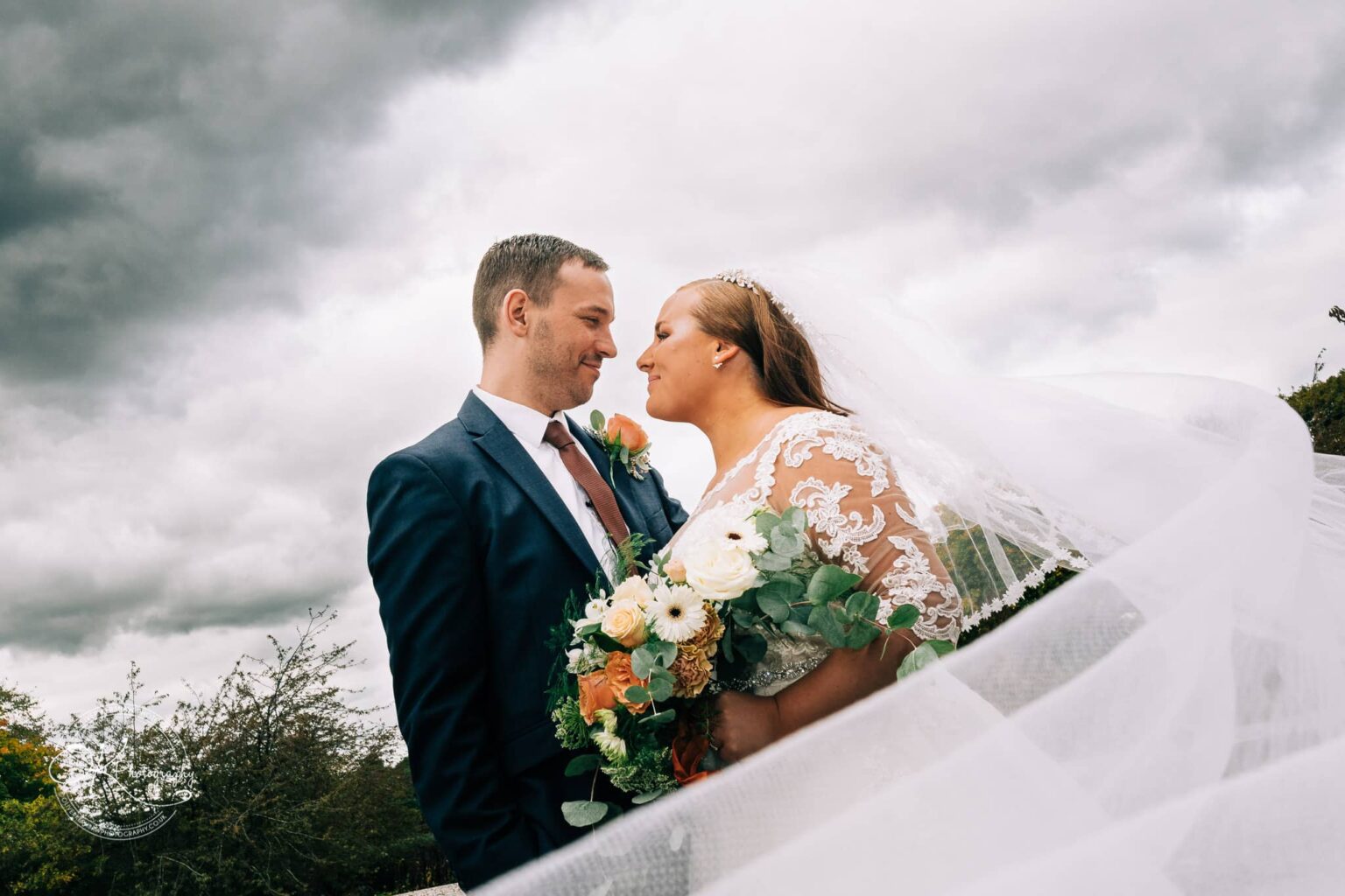 Wedding photography The Embankment Bride and groom smiling at each other under a cloudy sky, with the bride holding a bouquet of flowers and her veil flowing in the wind.