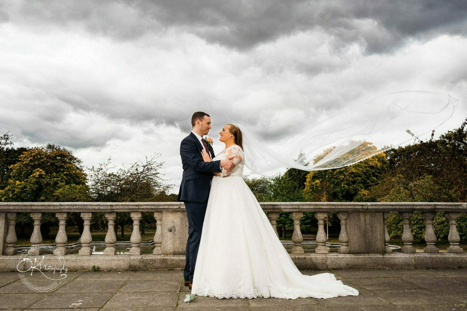 Wedding photography The Embankment A bride and groom embrace on a stone terrace with a dramatic, cloudy sky and trees in the background, the bride's veil flowing in the wind.