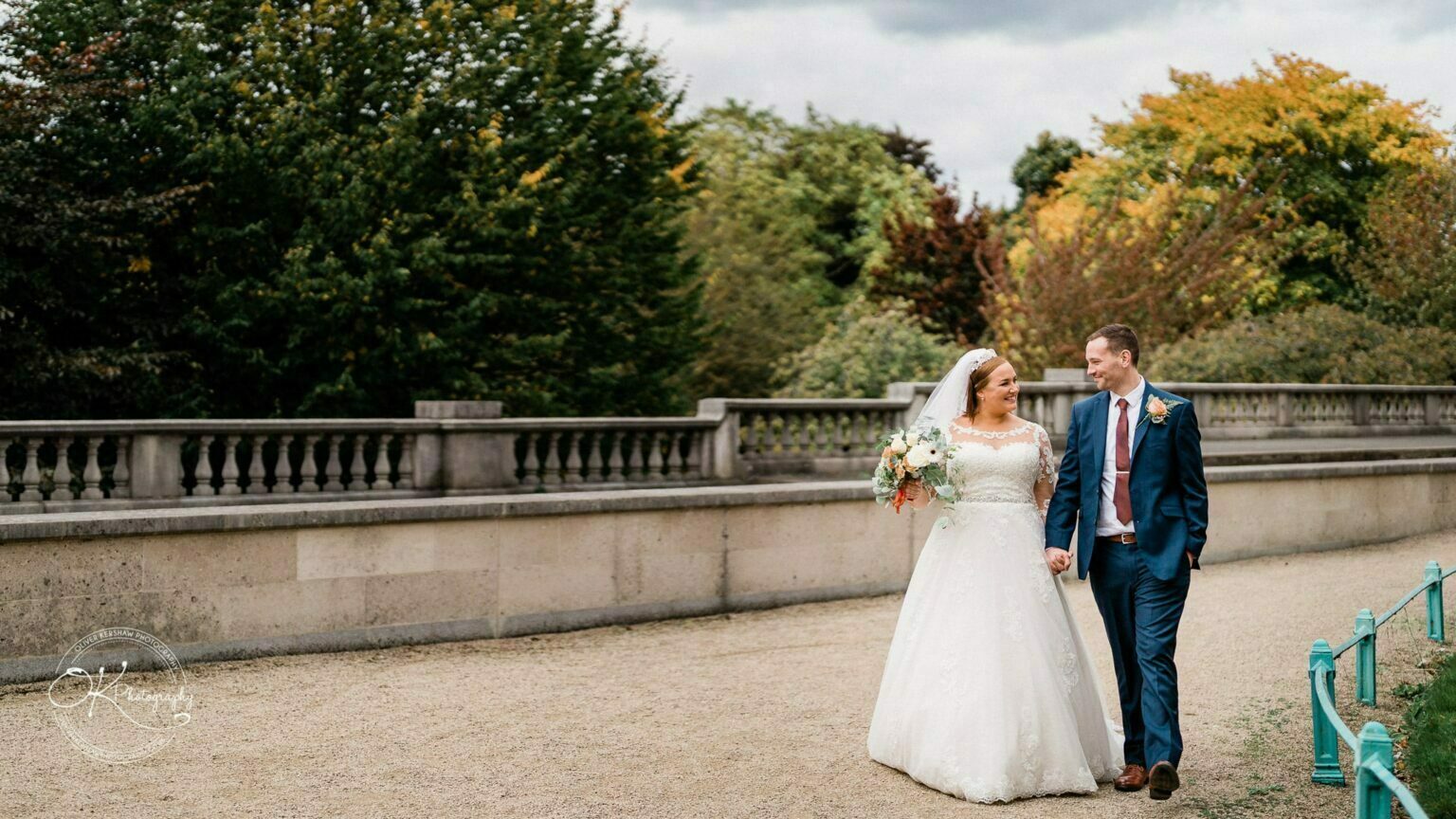Wedding photography The Embankment A bride and groom holding hands and walking along a path next to a stone balustrade, with trees and cloudy skies in the background.