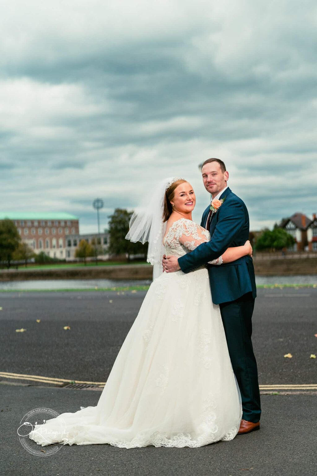 Wedding photography The Embankment Bride and groom embracing in wedding attire on a cloudy day, with buildings and trees in the background.