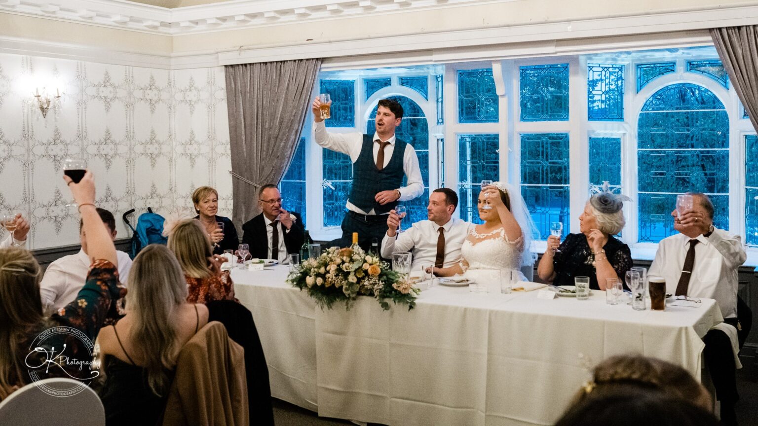Wedding photography The Embankment Wedding reception with guests raising glasses for a toast, including the bride in a white gown and a man giving a speech.