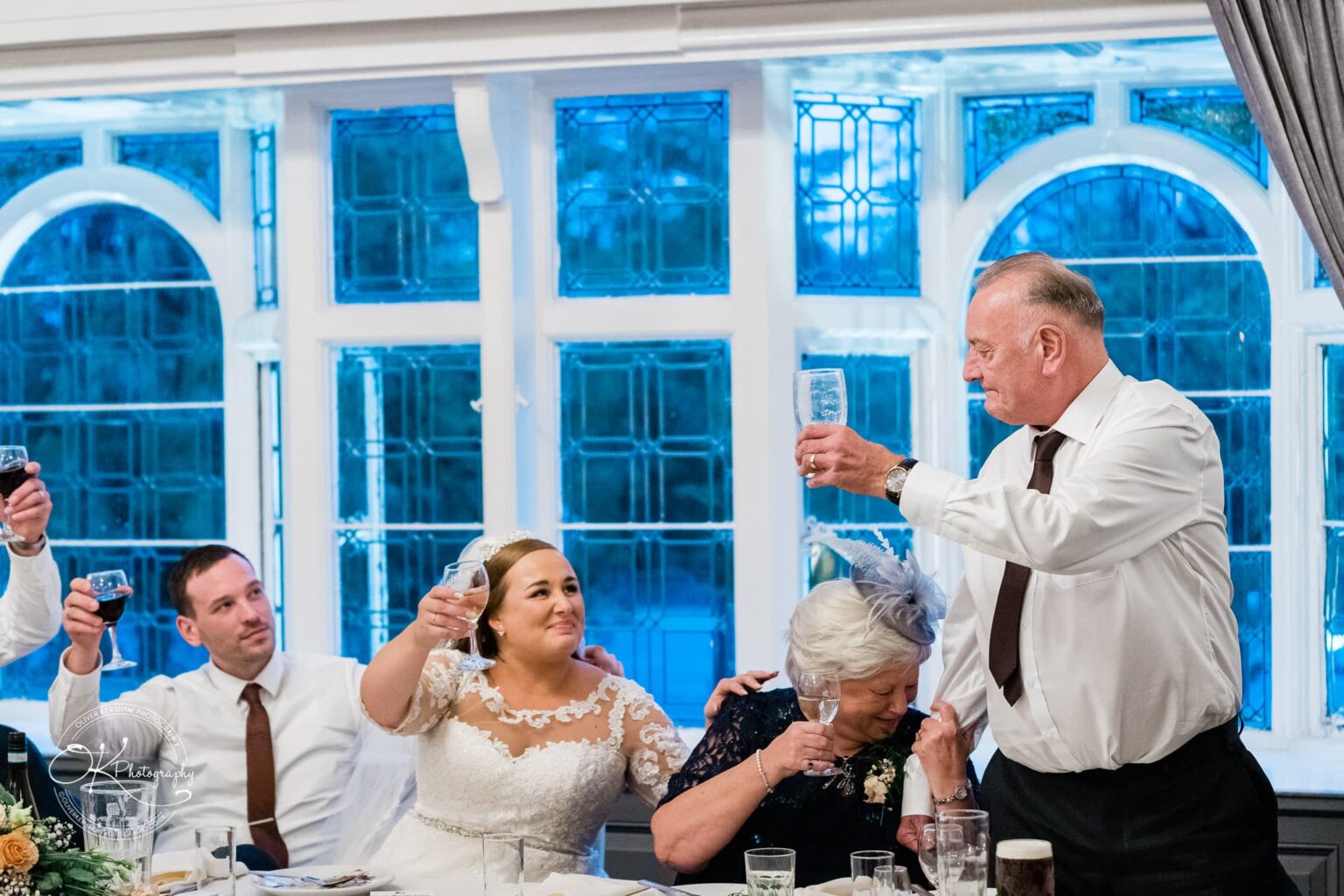 Wedding photography The Embankment Wedding party at a table raising their glasses for a toast, with blue stained-glass windows in the background.
