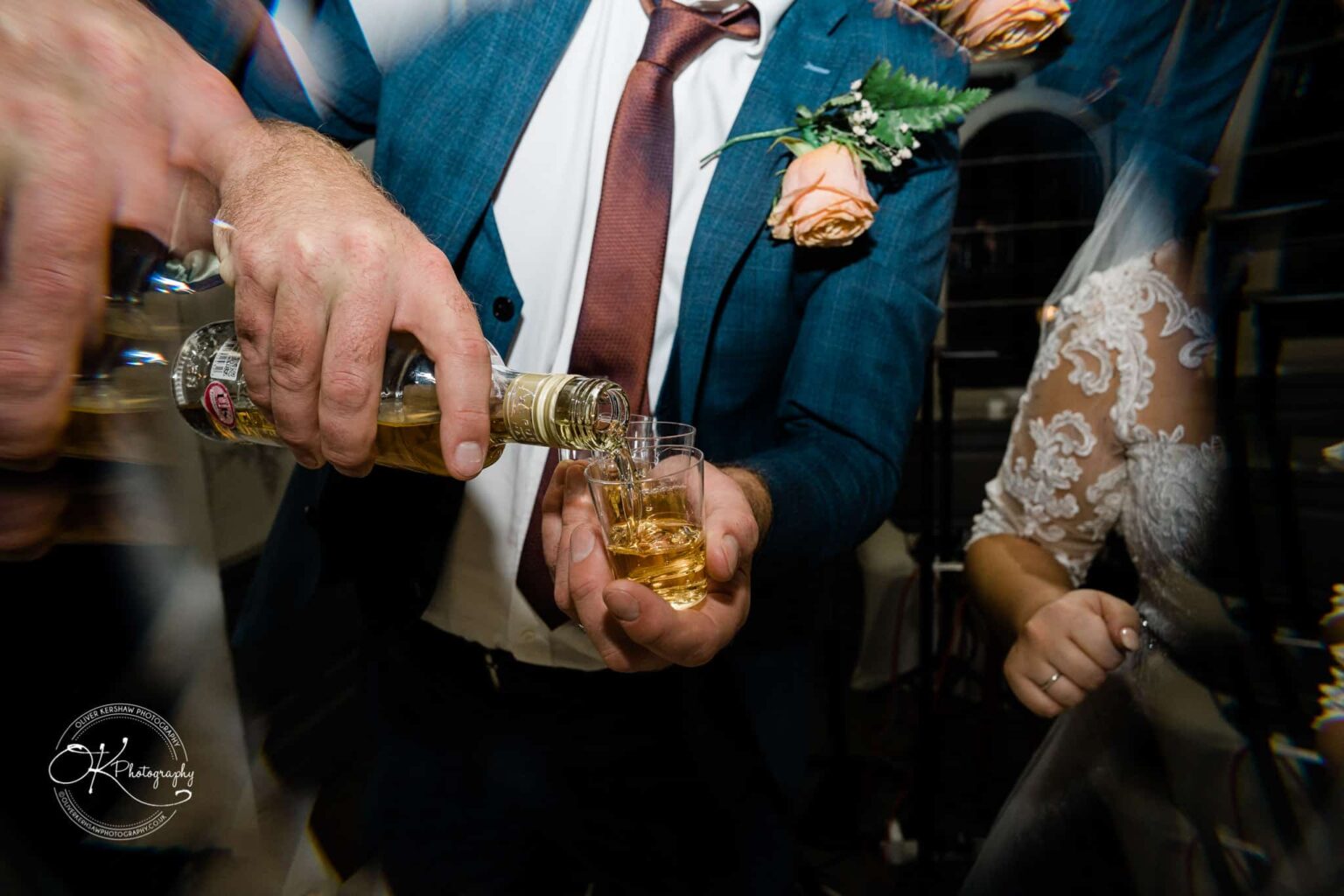 Wedding photography The Embankment A man in a blue suit pours liquor into a small glass, while a woman in a white lace dress stands beside him.