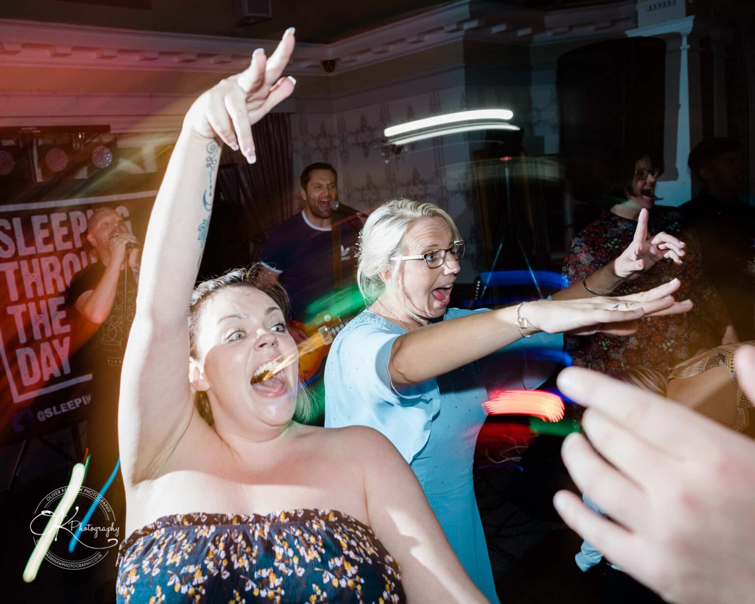Wedding photography The Embankment Two women enthusiastically dancing with raised arms in a lively, dimly lit venue with blurred colorful lights, and a music band performing in the background.