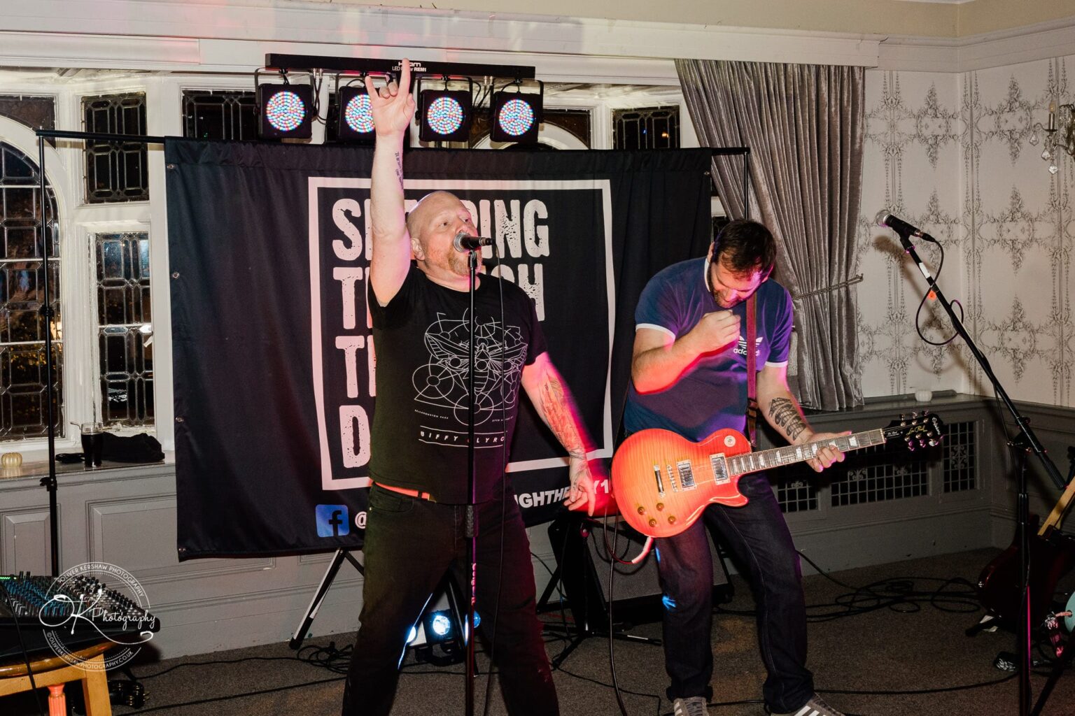 Wedding photography The Embankment Two musicians performing on stage, one singing with a microphone and raised arm, and the other playing an electric guitar, with a backdrop displaying "Singing Through The Dark."