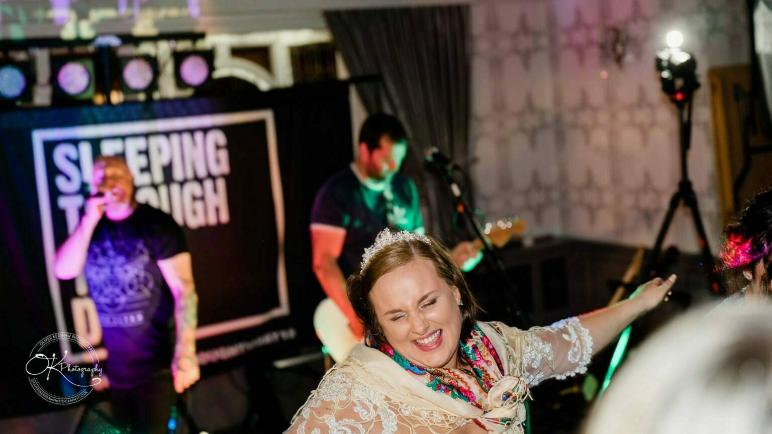 Wedding photography The Embankment A woman wearing a tiara and scarf dances in the foreground, while a band performs on stage in the background, with a sign reading "SLEEPING THROUGH" behind them.