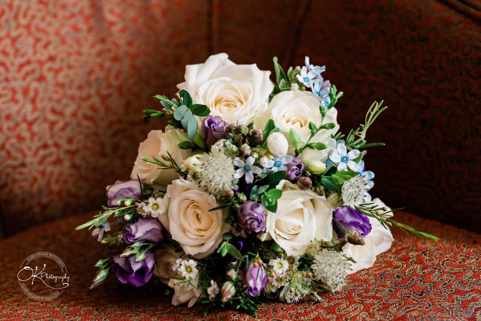 Yew Lodge Hotel Wedding Photography A bridal bouquet with white, purple, and blue flowers resting on a red patterned chair.