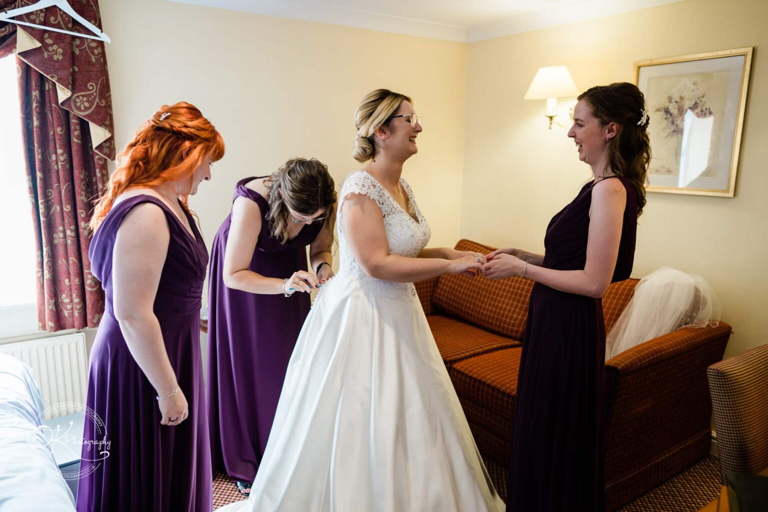 Yew Lodge Hotel Wedding Photography Bride in a white dress being assisted by bridesmaids in a hotel room.