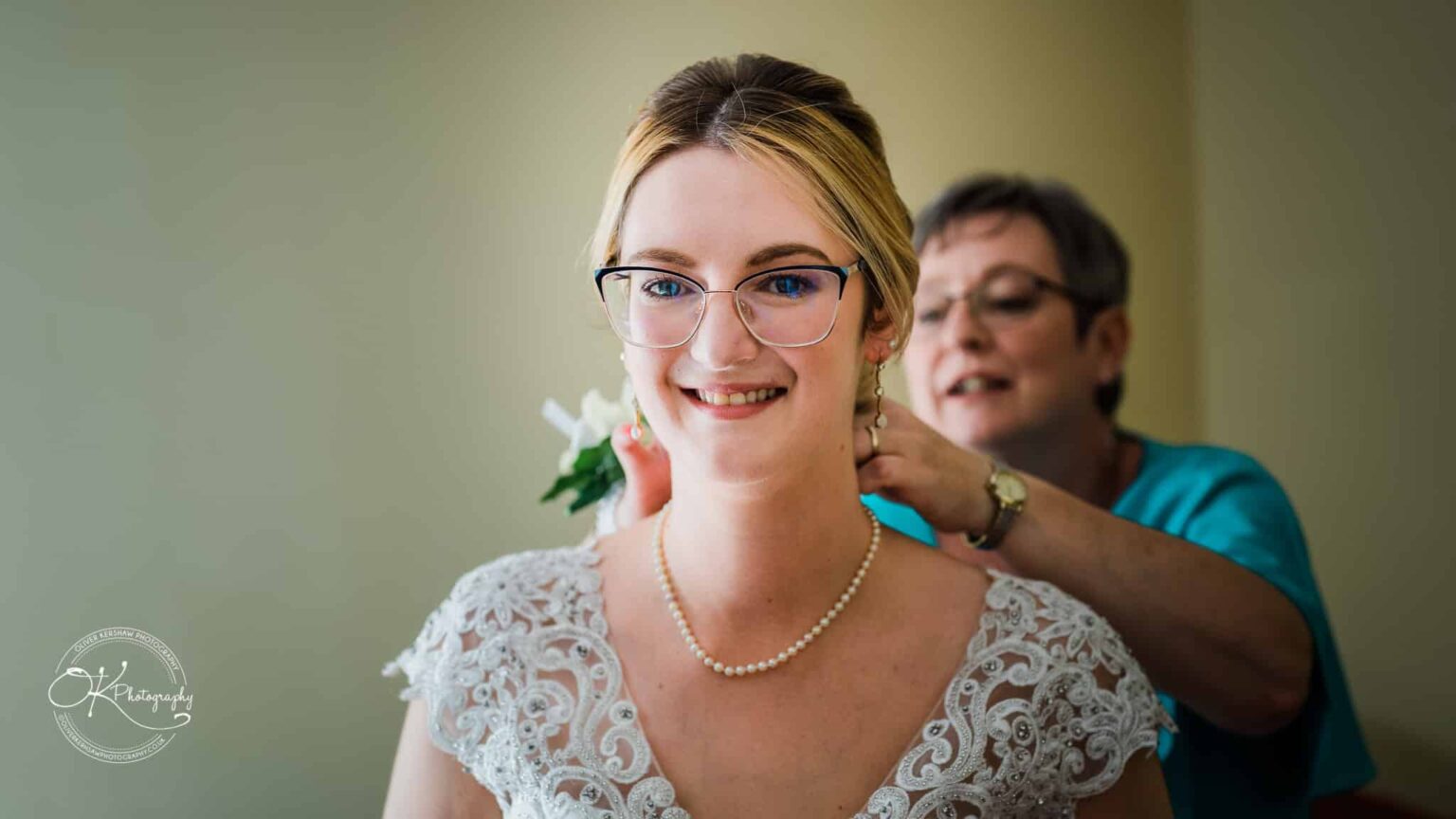 Yew Lodge Hotel Wedding Photography Bride smiling as a woman adjusts her necklace on her wedding day.