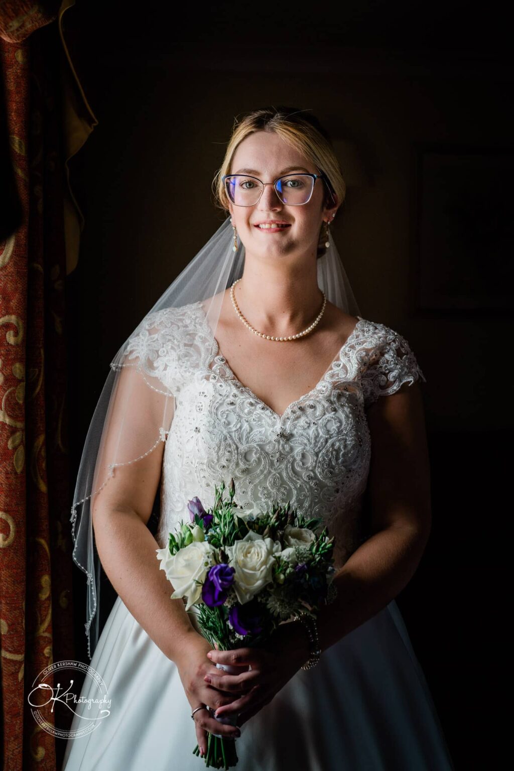 Yew Lodge Hotel Wedding Photography Bride with glasses and a lace wedding dress holding a bouquet of white and purple flowers, standing near a window.