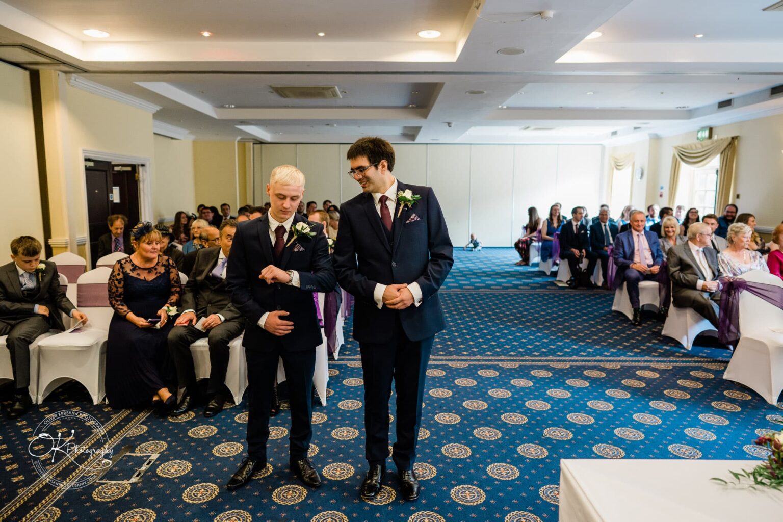 Yew Lodge Hotel Wedding Photography A wedding attendee checks his watch while standing next to another man in a suit, with seated guests in the background at Yew Lodge Hotel.