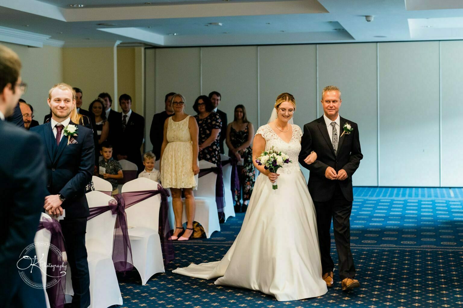 Yew Lodge Hotel Wedding Photography A bride in a white dress walks down the aisle with a man beside her, while guests look on and a groom in a suit waits at the front.