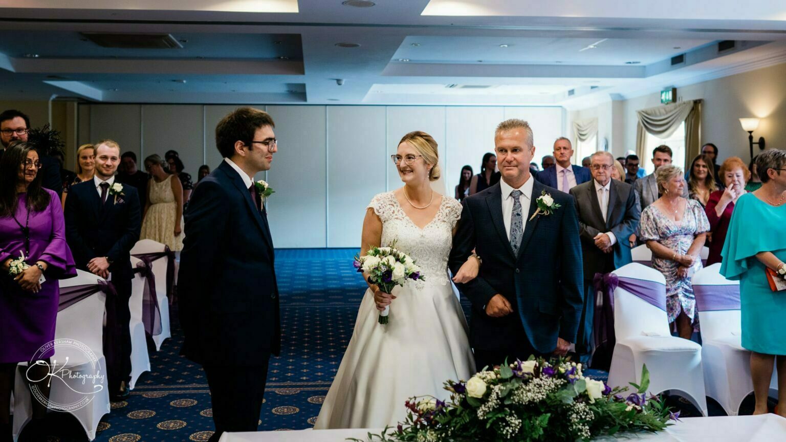 Yew Lodge Hotel Wedding Photography Bride and groom smiling at each other during wedding ceremony with guests seated behind them.